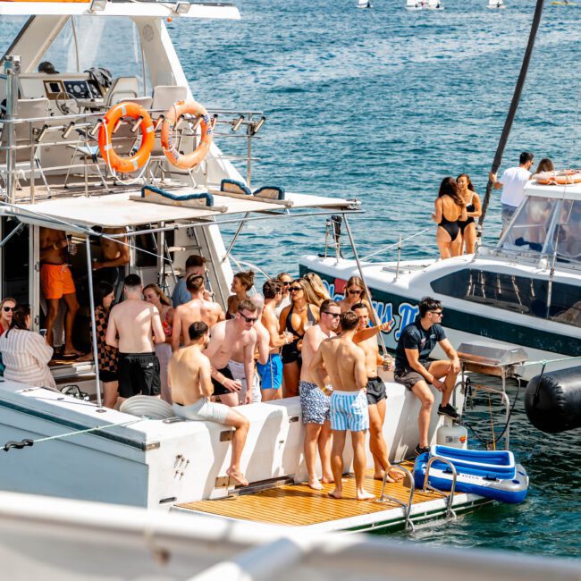 A group of people in swimwear socialize on the deck of a large yacht on the water, with another smaller boat nearby. The background features a cityscape and a partly cloudy sky, along with multiple sailboats on the horizon. It's clearly an event by The Yacht Social Club.