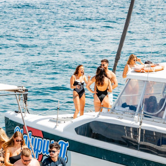 A group enjoys a sunny day on a boat. Five individuals stand on the deck, talking and taking photos, with one person adjusting a strap on their swimsuit. The calm blue waters of Sydney Harbour surround them, and other boats and swimmers are visible in the background. A perfect day for The Yacht Social Club Event Boat Charters!