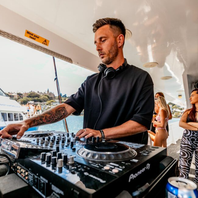 A DJ is immersed in his music, operating a mixer on a boat. The scene is lively with people in summer attire enjoying themselves. The surroundings include a body of water and distant trees, suggesting a warm, sunny day. Experience the vibe with The Yacht Social Club Sydney Boat Hire.
