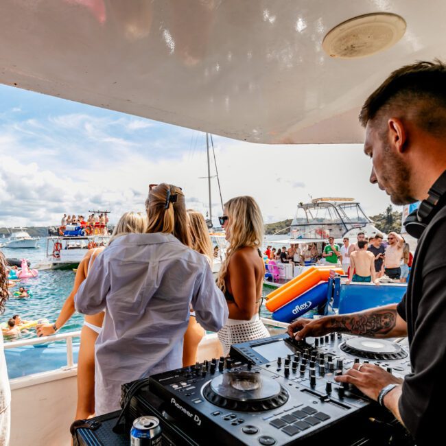 A DJ is playing music on a luxury yacht deck while a crowd enjoys a sunny day, some lounging on inflatables in the water and others dancing on the boat. The scene features a vibrant social gathering with clear skies and picturesque waters, courtesy of The Yacht Social Club Event Boat Charters.