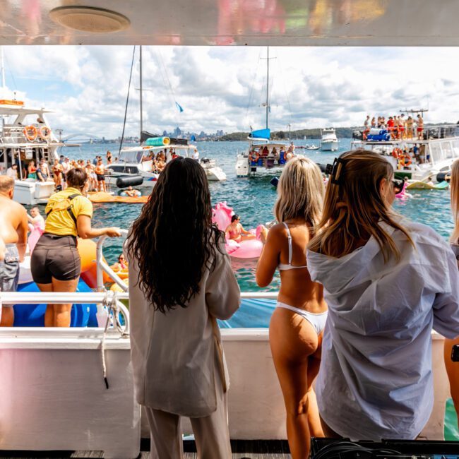 A group of people in swimwear and casual clothing enjoy a sunny day on a boat, with several other boats and people visible in the water and background. The scene suggests a lively and social atmosphere on a clear, bright day, perfect for Luxury Yacht Rentals Sydney.