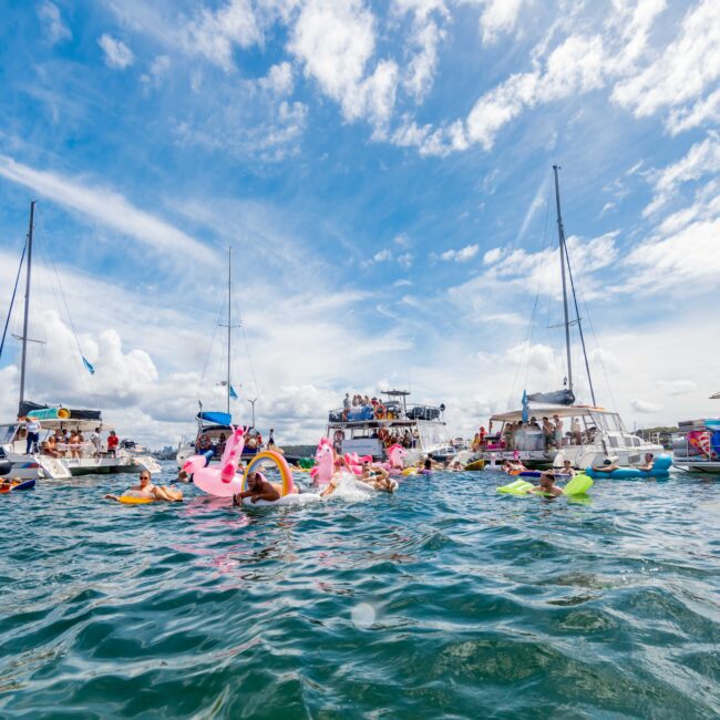 People are having a fun day in the water surrounded by boats. Some wear colorful floats shaped like flamingos and unicorns. The sky is dotted with white clouds, and some people are on the boats, socializing and enjoying the sun. The atmosphere is lively and festive, much like an event organized by The Yacht Social Club.