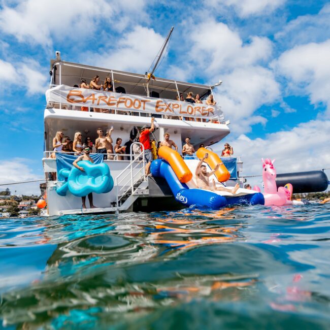 A double-decker boat named "Barefoot Explorer" is anchored with people on board enjoying the water. Large inflatable pool toys, including a blue horse and pink unicorn, float nearby. The sky is bright blue with scattered clouds, and people are waving and smiling at The Yacht Social Club event.
