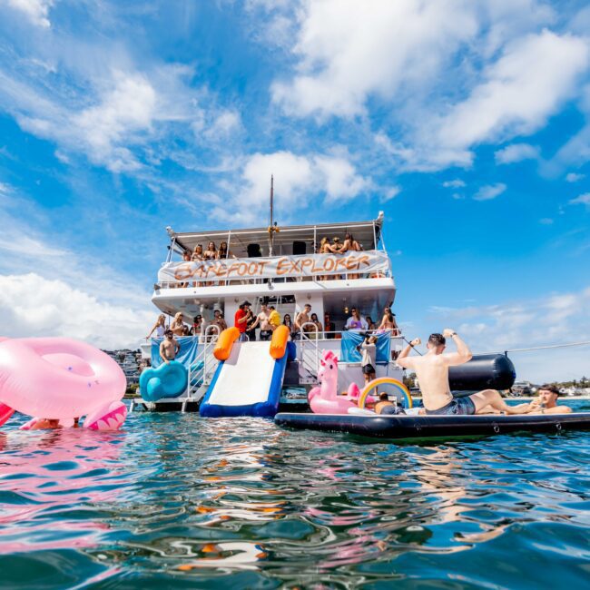 A group of people enjoying a sunny day on a luxury yacht rental with slides and inflatables in the water. Some are on top of the boat, while others swim and use the slide. The sky is blue with scattered clouds, perfect for a Yacht Social Club Sydney Boat Hire experience.