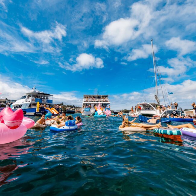 People relaxing on colorful inflatables in the water, surrounded by various boats. The sky is partly cloudy, and the water is calm. A festive atmosphere with participants enjoying a sunny day on a large body of water, hosted by Boat Parties Sydney The Yacht Social Club.