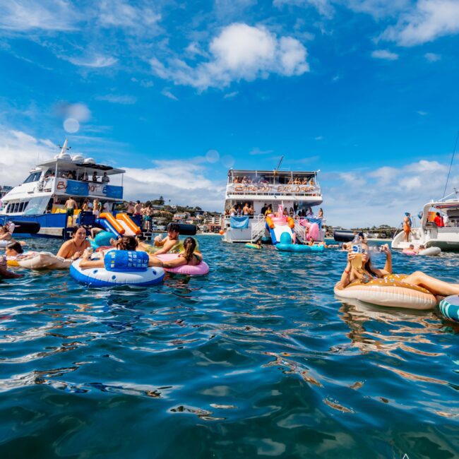 A lively scene of people enjoying a sunny day on the water, floating on inflatables and swimming near anchored boats in Sydney Harbour. The clear blue sky adds to the vibrant atmosphere. The Yacht Social Club logo is visible in the bottom right corner, showcasing their Boat Rental and Parties Sydney services.
