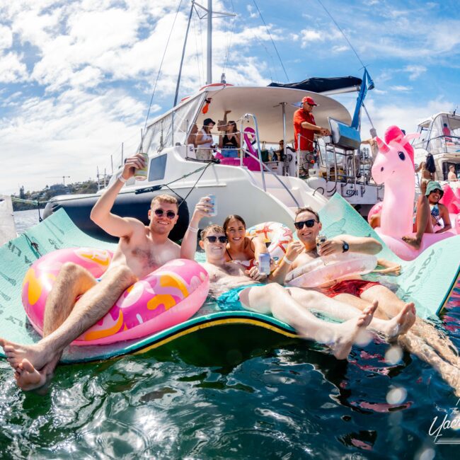 A group of five people lounges on an oversized inflatable mat in the water, next to a boat from Sydney Harbour Boat Hire. They hold drinks and smile at the camera. In the background, more people are on the boat with an inflatable pink unicorn under a clear sunny sky.