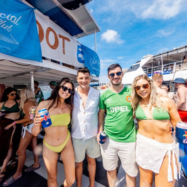 Four people are smiling and posing for a photo on a boat during The Yacht Social Club Sydney Boat Hire party. Two women in swimsuits stand beside two men—one in a white shirt and the other wearing a green shirt with "Opel" written on it. They are holding drinks, with a lively crowd in the background.