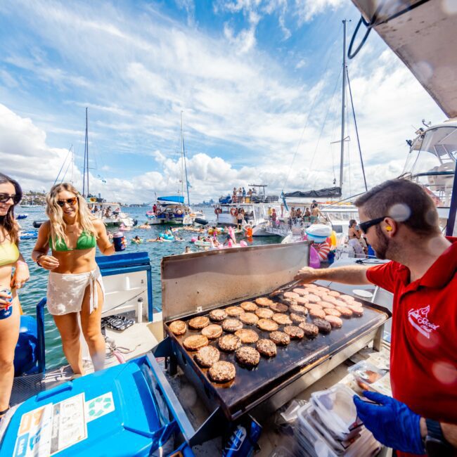 A man in a red shirt cooks burgers on a grill attached to a boat while two women in swimsuits stand nearby. In the background, people are swimming and enjoying themselves amidst other boats. The sunny setting with partly cloudy skies depicts a festive atmosphere, perfect for The Yacht Social Club Sydney Boat Hire.
