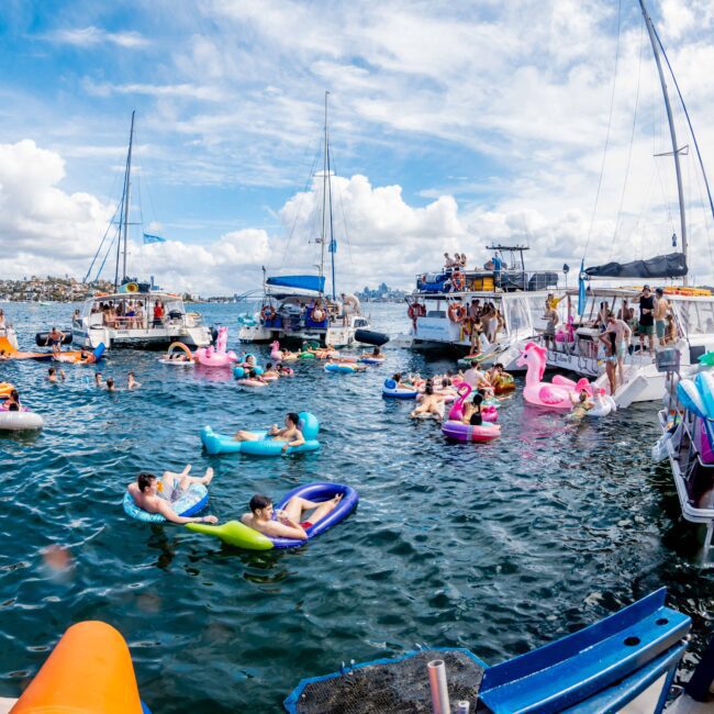 People are enjoying a sunny day at The Yacht Social Club Sydney Boat Hire party with multiple boats and colorful inflatables in the water. Some individuals are floating on inflatables shaped like flamingos and unicorns, while others are swimming or socializing on the decks of various boats.