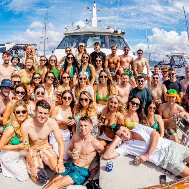 A large group of people wearing sunglasses and summer clothing are gathered on the deck of a boat, smiling and posing for a photo during The Yacht Social Club event. The sky is blue with some clouds, and the water is visible in the background. Some are holding drinks, and one person has a camera.