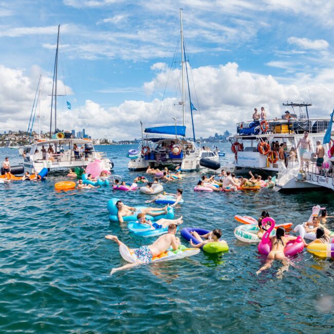 A lively scene on a sunny day with people on various colorful inflatable floats in the water. Boats from The Yacht Social Club are docked nearby, with more people on board, enjoying the weather. The sky is filled with fluffy clouds, adding to the cheerful ambiance.