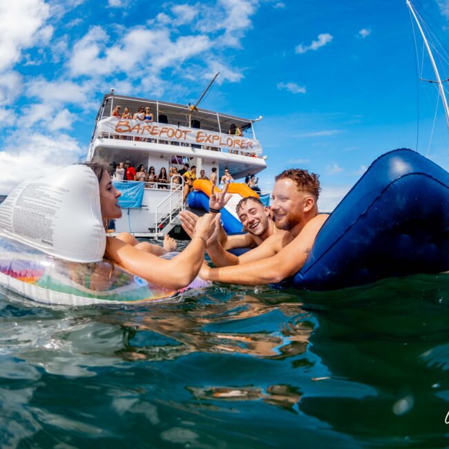 A group of people enjoying a day on the water, with three individuals arm wrestling on inflatable rafts. In the background, a boat named "Barefoot Explorer" is docked, filled with others engaged in leisure activities. Hosted by The Yacht Social Club, the bright blue sky and calm water set the perfect scene.