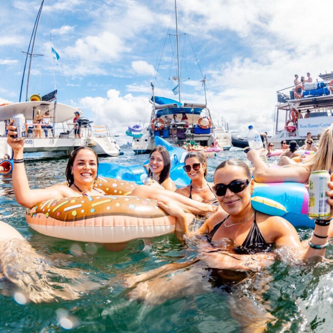 A group of people are enjoying a sunny day in the ocean, lounging on inflatable floats and holding drinks. Boats and yachts from The Yacht Social Club Sydney Boat Hire can be seen in the background, along with other people in the water. The sky is blue with a few clouds.
