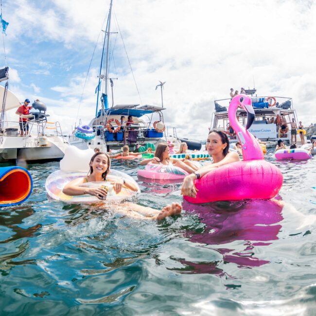 People are enjoying a fun time on inflatable floats and boats in a lively marina setting. Some are floating on unicorn, flamingo, and donut-shaped floats, while others relax on nearby boats. The sky is partly cloudy, creating a cheerful, vacation-like atmosphere. This could be your next adventure with The Yacht Social Club Sydney Boat Hire.