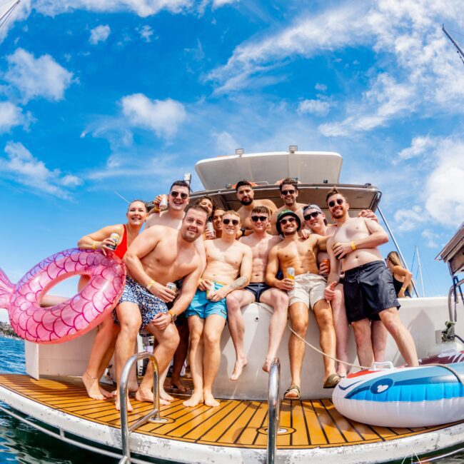 A group of eleven men and one woman pose together on the deck of a boat under a bright blue sky. They are in swimwear, and some are holding inflatable pool toys, including a pink mermaid tail. The group is smiling and appears to be enjoying a sunny day on the water with Sydney Harbour Boat Hire The Yacht Social Club.