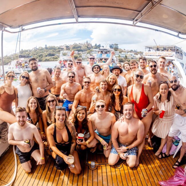 A large group of people, wearing swimsuits and summer clothing, stand closely together on a boat, smiling and holding drinks. More boats and people are visible in the background, creating a lively and festive atmosphere on the water at The Yacht Social Club.