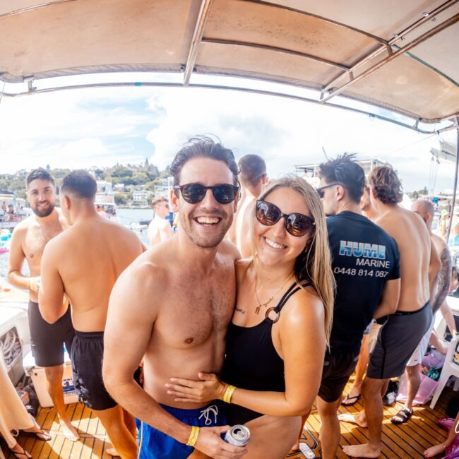 A shirtless man and a woman in sunglasses smile at the camera while embracing on a boat crowded with people. The woman is wearing a black swimsuit and holding a drink. Other partygoers are visible in the background, some wearing swimsuits and holding drinks, enjoying The Yacht Social Club event.