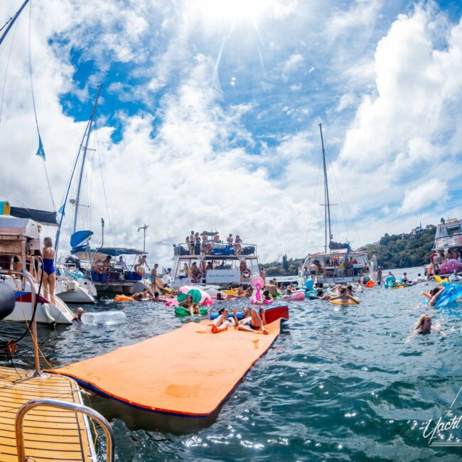 A lively scene with people swimming and lounging on inflatable floats. Boats and yachts from The Yacht Social Club are anchored nearby under a bright blue sky with scattered clouds. In the foreground, a large orange floating mat extends into the water. The atmosphere is festive and social, perfect for boat rental parties in Sydney.