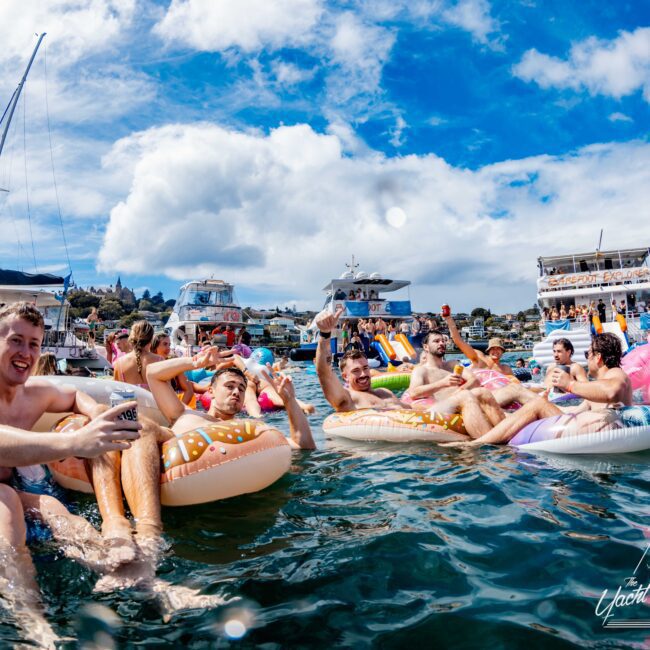 A joyful group lounges on colorful inflatable pool floats in the water, with yachts in the background under a partly cloudy sky. Participants, including some holding drinks, enjoy a sunny day. A logo in the corner reads "Boat Rental and Parties Sydney The Yacht Social Club.