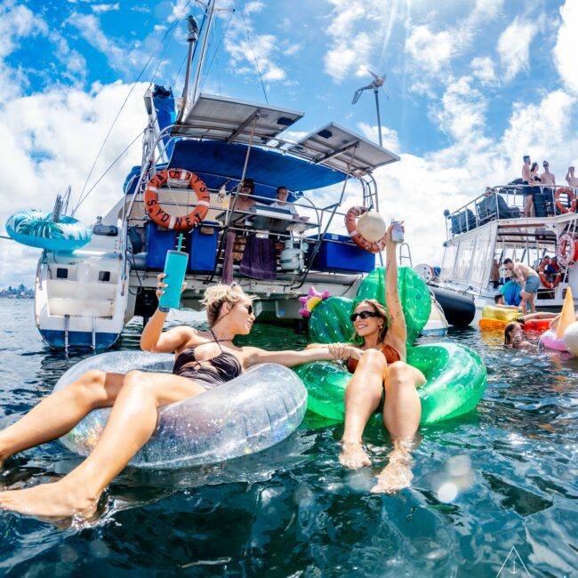Two women relax on inflatable floaties in the sea near a boat with people onboard. The sky is sunny with scattered clouds. One woman holds a drink while the other reaches out to touch her hand, creating a cheerful and fun atmosphere. The logo "The Yacht Social Club" is at the bottom right, highlighting Luxury Yacht Rentals Sydney.