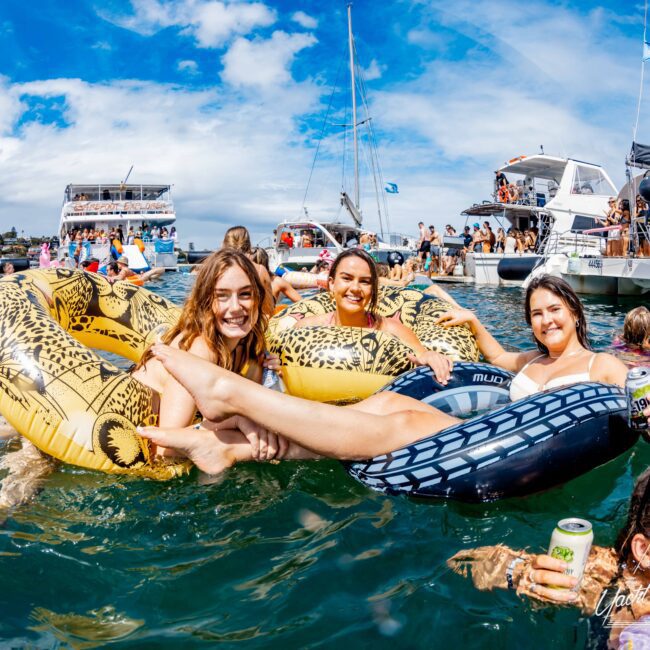 Three people smile while relaxing on inflatable tubes in the water, surrounded by boats and other people. Two hold drinks as they enjoy a sunny day with clear skies. The lively scene features The Yacht Social Club Sydney Boat Hire offering luxury experiences and fun activities in the background.