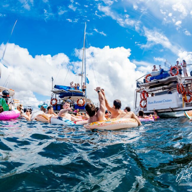 A vibrant scene of people floating on colorful inflatable tubes in the ocean, surrounding anchored boats. The clear blue sky is dotted with fluffy white clouds. The atmosphere is lively and joyful, with groups of friends engaging in summer fun on the water at The Yacht Social Club Sydney Boat Hire.