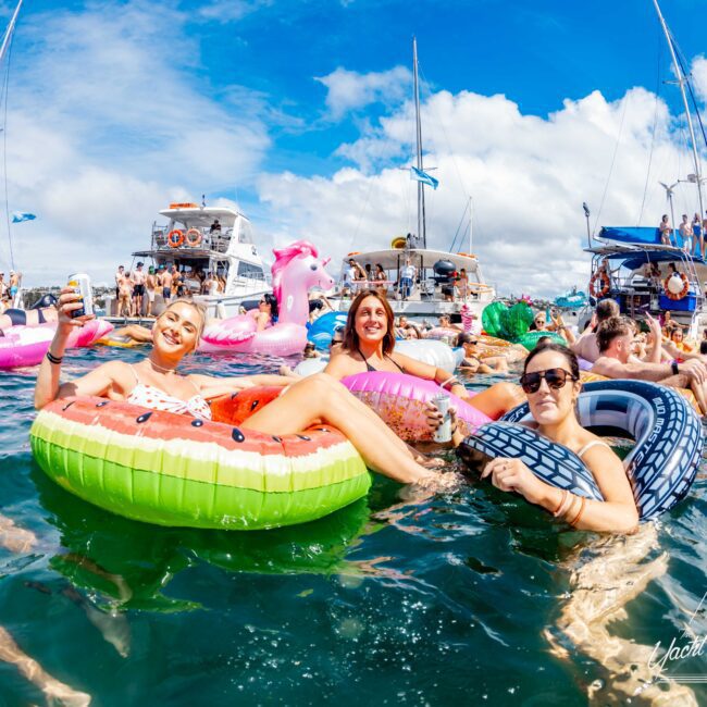 A group of people enjoying a sunny day on inflatable floats of various shapes in a body of water, surrounded by boats. Three women are prominently smiling and relaxing on water floats shaped like a watermelon slice, donut, and tire. Experience the bright and festive atmosphere with Sydney Harbour Boat Hire The Yacht Social Club.
