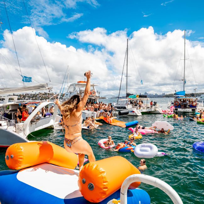 A lively scene of people celebrating on yachts and inflatables in the water. The central focus is a person about to jump from a large inflatable platform into the water. The sky is partly cloudy, and the atmosphere is energetic and fun at a Boat Parties Sydney by The Yacht Social Club.