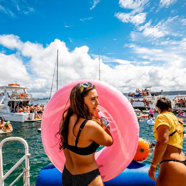 Two women in swimsuits navigate a large inflatable pink float on the deck of a boat. The surrounding water is crowded with boats and people enjoying the sunny day. The sky is partly cloudy, and several other boats and people are visible in the background, highlighting a lively scene from The Yacht Social Club Event Boat Charters.