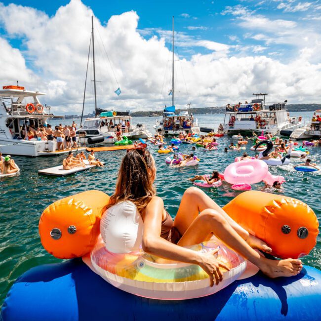 A woman lounges on a colorful inflatable float in a vibrant and busy ocean party scene. Numerous boats are anchored close together, with people enjoying the water on various floats and swimming. The sky is bright blue with scattered clouds, embodying the essence of Boat Parties Sydney The Yacht Social Club.