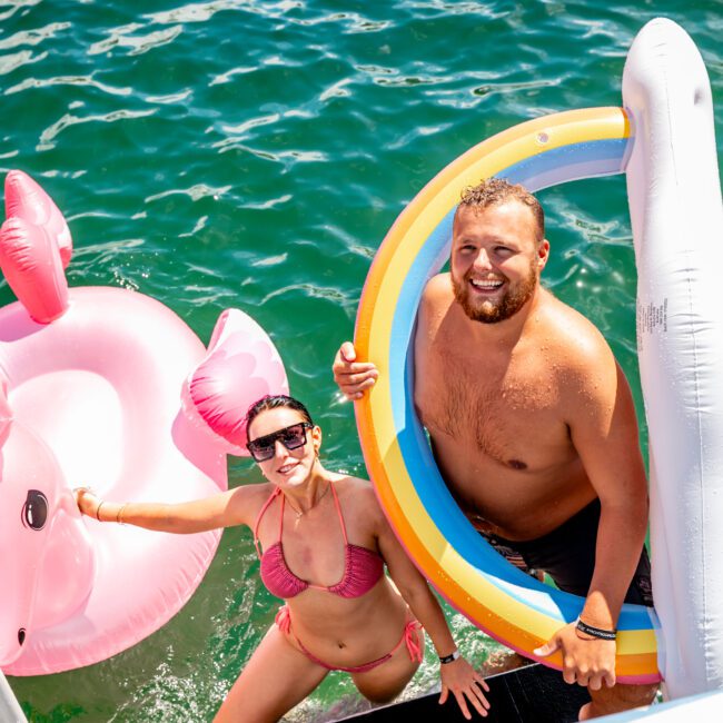 A man and a woman stand on a dock by the water. The man holds a colorful pool float, while the woman leans on a pink flamingo float. Both are in swimwear, smiling and enjoying the sunny weather. Behind them, luxury yachts from The Yacht Social Club Sydney Boat Hire are docked against the bright, clear water.