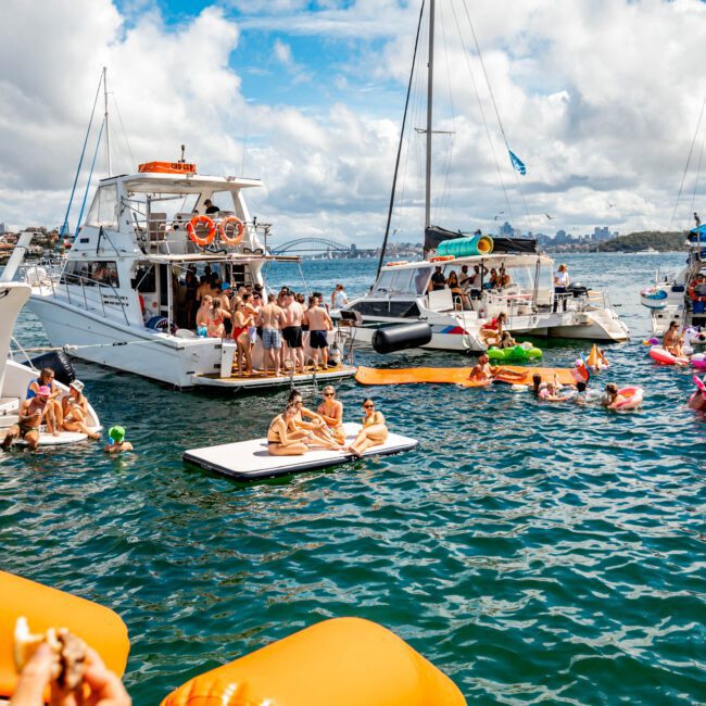 A lively scene of a boat party on a sunny day features several yachts and boats anchored close together. People are swimming, floating on inflatable tubes, and socializing. The backdrop includes a city skyline and a large bridge under a partly cloudy sky—perfect for The Yacht Social Club Sydney Boat Hire.