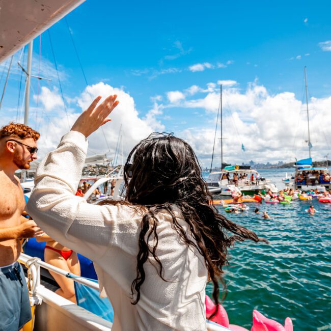 People aboard a boat are enjoying a sunny day by waving and observing others in the water. The scene features colorful inflatable floats and multiple boats, including luxury yacht rentals, anchored in a scenic location. The water sparkles under a bright blue sky with scattered clouds.