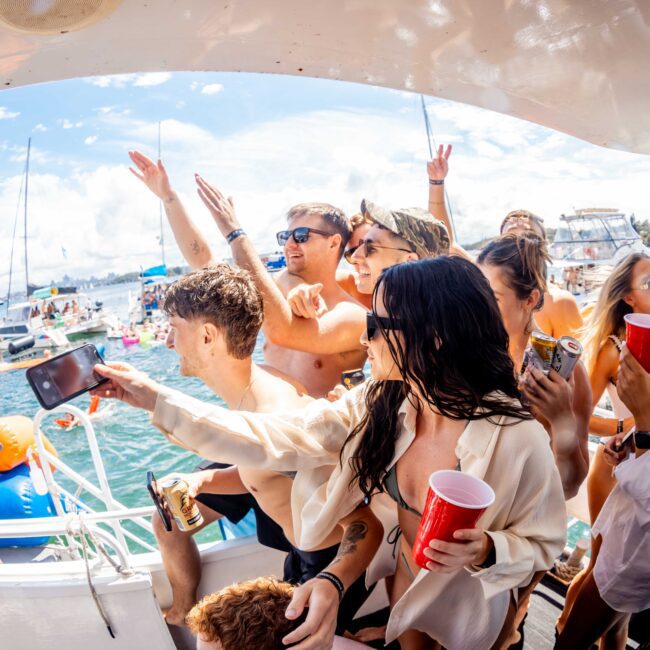 A group of people are having fun on a boat during a sunny day. They are holding red cups, wearing casual summer attire, smiling, and taking photos. The Yacht Social Club Sydney Boat Hire provides an ideal backdrop with other boats on the water and a partly cloudy sky.