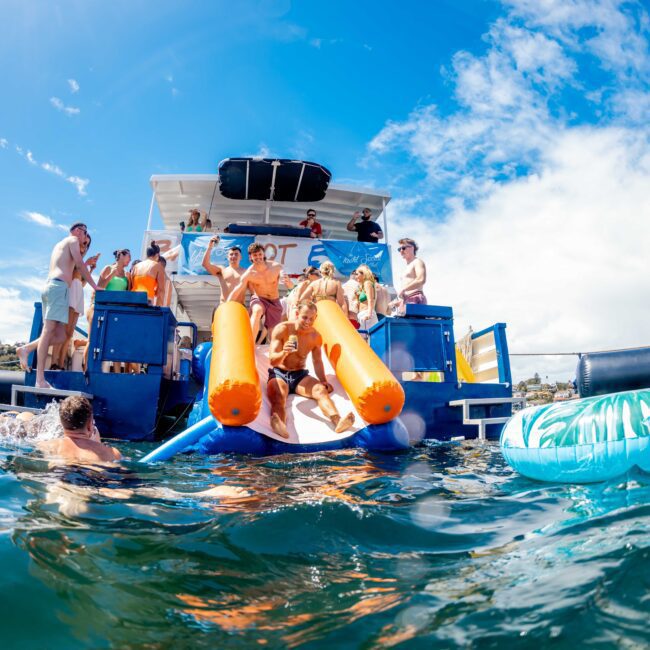 People are enjoying a sunny day on and around a yacht with an inflatable slide. Some individuals are in the water with inflatables, while others relax on the deck. The clear blue sky and water create a vibrant, fun atmosphere, perfect for Boat Parties Sydney The Yacht Social Club.