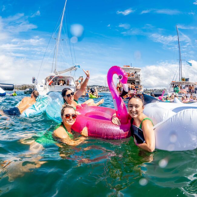 A group of people enjoying a lively boat party in clear blue waters. They smile and pose with colorful inflatable floats, including a pink flamingo and a white swan. Nearby yachts and a slide enhance the festive atmosphere under a bright, sunny sky hosted by The Yacht Social Club Sydney Boat Hire.