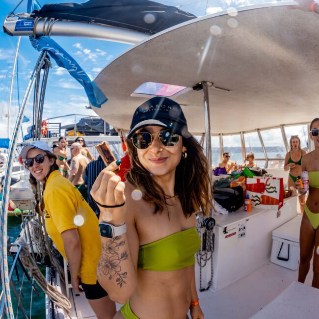 People on a boat enjoy a sunny day. A woman in a green bikini and cap holds a snack, smiling. Others, also in swimsuits, are socializing. Various boats and inflatables are in the background on the water. The mood is festive and lively, capturing the essence of The Yacht Social Club's Boat Parties Sydney.
