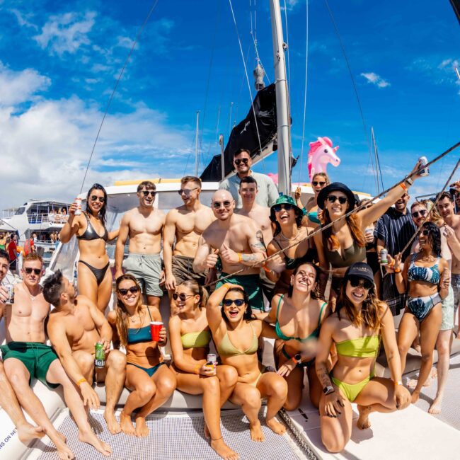 A large group of people wearing swimsuits and sunglasses standing on the deck of a boat with clear blue skies overhead. They appear to be celebrating and having a good time, with some holding drinks and posing cheerfully for the photo, likely enjoying The Yacht Social Club Sydney Boat Hire experience.