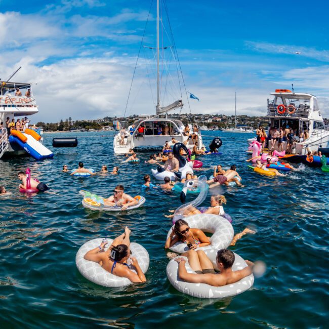 A lively scene of people floating on various inflatables, including a swan and a flamingo, in blue-green water near anchored boats. The Yacht Social Club Sydney Boat Hire logo is visible in the bottom right corner of the image against a partly cloudy sky.