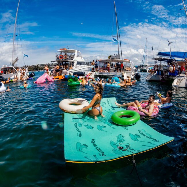 People are enjoying a sunny day on the water, relaxing on various floats and inflatable mats surrounded by yachts. The scene, part of The Yacht Social Club Sydney Boat Hire, is vibrant with activity as individuals socialize and sunbathe. The sky is partly cloudy, and the atmosphere is lively and festive.