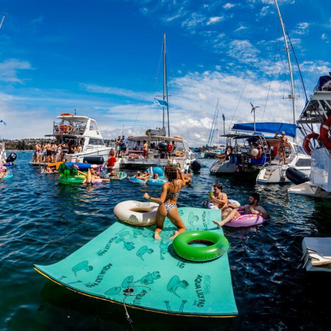 A lively scene of people enjoying a sunny day on the water, surrounded by boats. Individuals are lounging on colorful floaties and a large floating mat, socializing and soaking up the sun. Sailboats and yachts from The Yacht Social Club Sydney Boat Hire are anchored nearby under a clear blue sky.