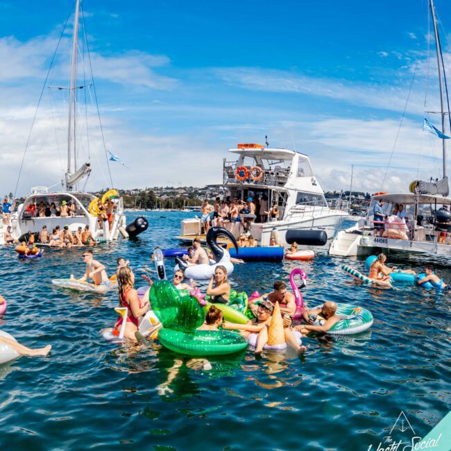 A lively party scene on the water with numerous people on inflatables, including a unicorn and a cactus, surrounded by several boats. The sky is clear, and the mood is festive as people enjoy the sunny day. The Yacht Social Club Sydney Boat Hire logo is in the bottom right.