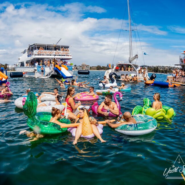A lively pool party scene with people enjoying themselves on various inflatable floats in the water. Boats are docked nearby, and some guests are standing or sitting on the decks. The atmosphere is festive, with a bright, sunny day enhancing the fun at The Yacht Social Club Event Boat Charters.