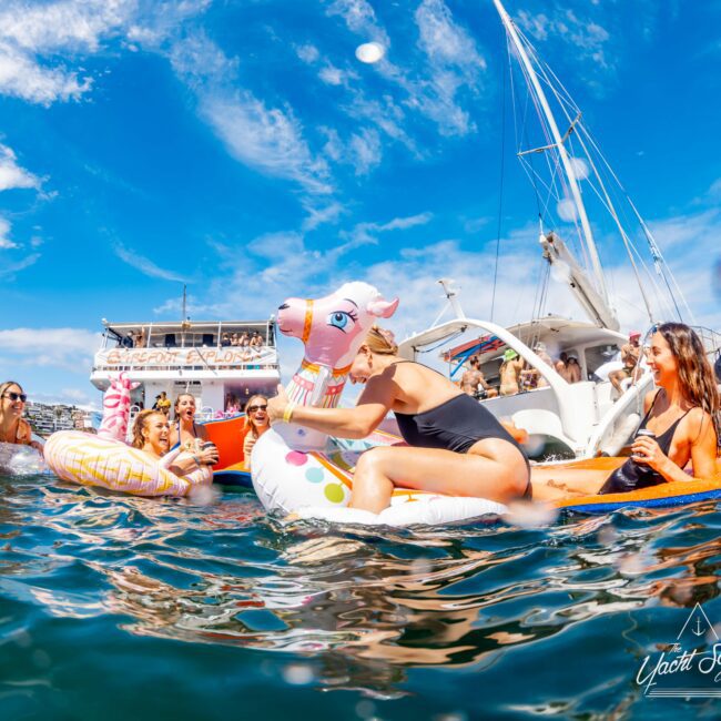 A group of people enjoying a sunny day on the water, surrounded by boats. Some are on pool floats, including a unicorn and donut float. They are laughing and engaging in a lively, fun atmosphere. The sky is clear with some clouds. The logo "The Yacht Social Club Sydney Boat Hire" is visible.