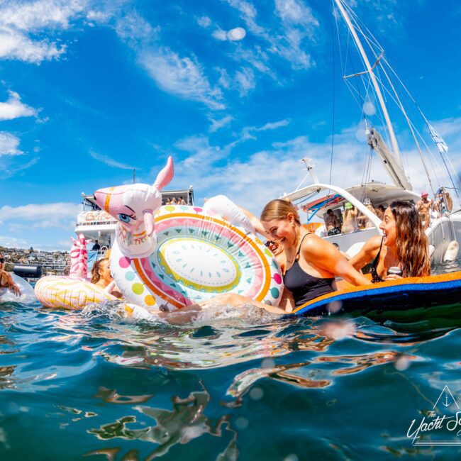 A group of smiling people enjoy a sunny day on the water, holding onto an inflatable unicorn float. Boats line the background under a blue sky with scattered clouds. The water is clear, and everyone appears happy and relaxed. The Yacht Social Club logo is visible in the corner, showcasing Sydney Harbour Boat Hire The Yacht Social Club.