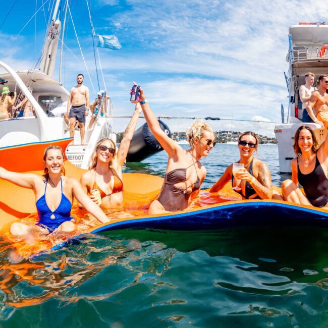 A group of five women in swimsuits relax and enjoy drinks on an orange floating mat in the water, surrounded by boats and other people. The sunny scene, part of a Boat Rental and Parties Sydney The Yacht Social Club event, shows a lively atmosphere with blue skies and others enjoying the summer day on boats in the background.