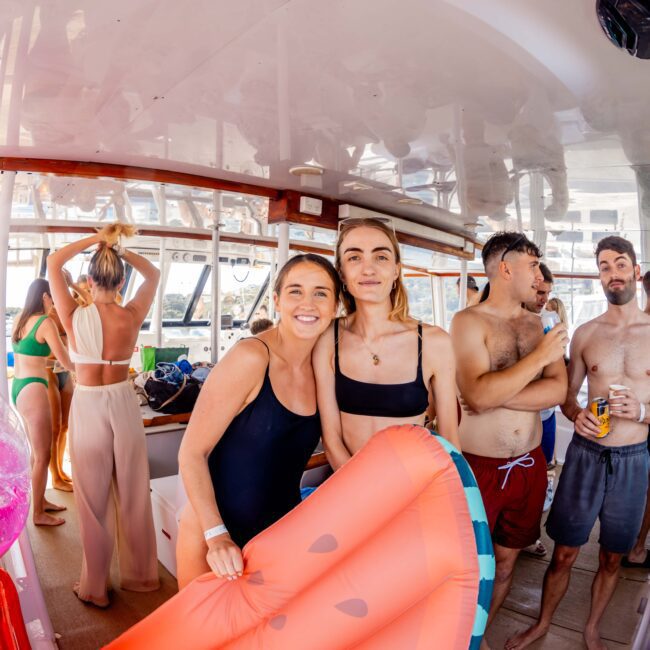 A group of people enjoying a boat party, some in swimsuits, standing and sitting around inside the boat from The Yacht Social Club Sydney Boat Hire. Two women in the center pose with a large inflatable watermelon slice. A pink flamingo float is also visible on the left. The boat's interior is bright and lively.