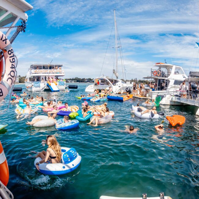 A lively scene of people enjoying a sunny day on the water, floating on inflatables and surrounded by boats. The festive atmosphere is boosted by Sydney Harbour Boat Hire The Yacht Social Club. Various colorful floats and enthusiastic individuals create a vibrant setting under a clear sky with some white clouds. A life ring is visible in the foreground.