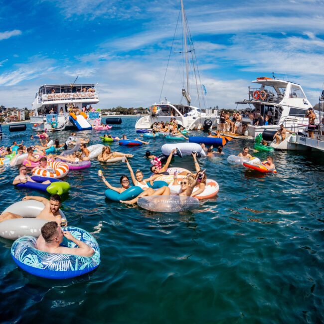 A lively scene of people enjoying a sunny day on the water, floating on various inflatables near several yachts. The water is a clear blue, and the sky is partly cloudy. Everyone appears to be having a great time socializing and relaxing at The Yacht Social Club Event Boat Charters. "Yacht Social Club" logo visible.